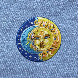 May include: A circular embroidered patch featuring a yellow sun with a face and a blue crescent moon. The design includes small white stars on a dark blue background, with golden rays extending from the sun, all stitched onto blue denim fabric.