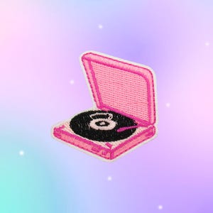 May include: An embroidered patch featuring a pink portable record player with an open lid. The device has a black circular vinyl record in the center with white details, and a pink tone arm positioned over the record, rendered in stitched thread.