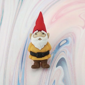 May include: A small, yellow gnome figurine with a red hat and white beard. The gnome is standing on a white and pink marbled surface.