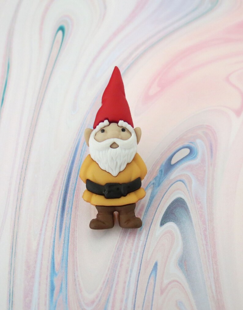 May include: A small, yellow gnome figurine with a red hat and white beard. The gnome is standing on a white and pink marbled surface.