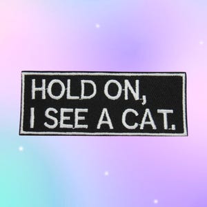 May include: A rectangular embroidered patch with a black background and a white border. The text HOLD ON, I SEE A CAT. is stitched in white capital letters across the center of the patch, creating a clear contrast against the dark fabric base.