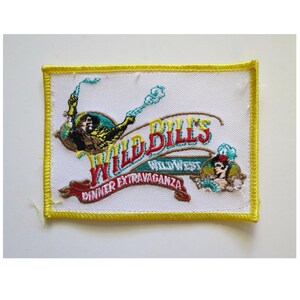 May include: Embroidered patch with a yellow border and a white background. The patch features the text "Wild Bill's Wild West Dinner Extravaganza" in red, teal, and gold. Illustrations of a cowboy and a woman are also included.