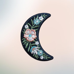 May include: A crescent moon-shaped embroidered patch featuring a dark navy blue background decorated with a variety of stitched flowers in shades of light pink, white, and bright blue, accented by green leaves and stems throughout the design.