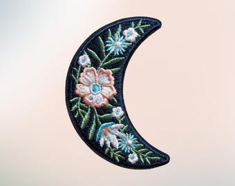 May include: A crescent moon-shaped embroidered patch featuring a dark navy blue background decorated with a variety of stitched flowers in shades of light pink, white, and bright blue, accented by green leaves and stems throughout the design.