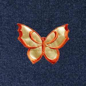 May include: A butterfly-shaped patch featuring a shiny cream-colored center with a vibrant orange embroidered border, securely attached to a dark blue denim fabric background with a visible diagonal twill weave texture throughout the material.