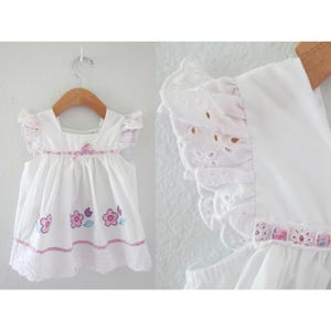 May include: White baby dress with floral embroidery and eyelet lace trim. The sleeveless dress features a square neckline, a pink ribbon, and a scalloped hem. The dress is hanging on a wooden hanger.