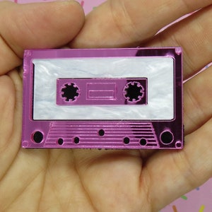 May include: A pink cassette tape charm with a white cassette tape inside. The charm is made of acrylic and has a shiny finish.