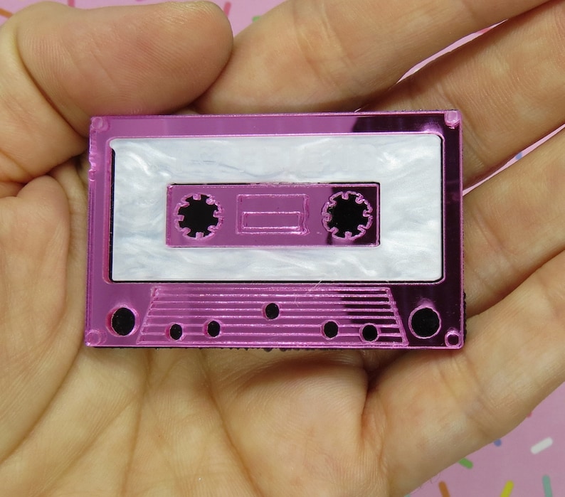 May include: A pink cassette tape charm with a white cassette tape inside. The charm is made of acrylic and has a shiny finish.