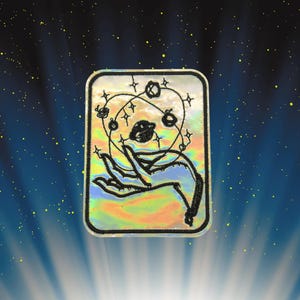 May include: A rectangular embroidered patch with a holographic, iridescent surface. The design features a black line art illustration of a hand holding a cluster of orbiting planets and stars, all enclosed within a thick black stitched border.