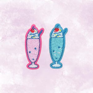 May include: Two embroidered patches shaped like milkshakes. One is pink with a pink outline, the other is blue with a blue outline. Each has a white whipped cream top with a red cherry and a blue straw.