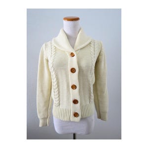 May include: A cream-colored knit cardigan featuring a shawl collar, long sleeves, and five brown buttons down the front. The garment displays a textured stitch pattern on the body and vertical cable knit detailing along the front panels.