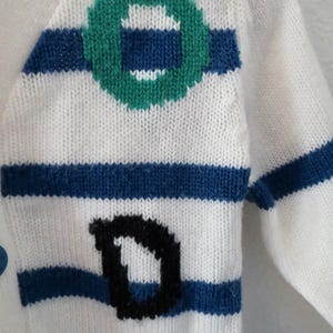 May include: A white knit cardigan featuring horizontal blue stripes, a green circular graphic, and a black letter D. The garment includes blue buttons along the front edge, showcasing a textured knit pattern throughout the fabric construction.