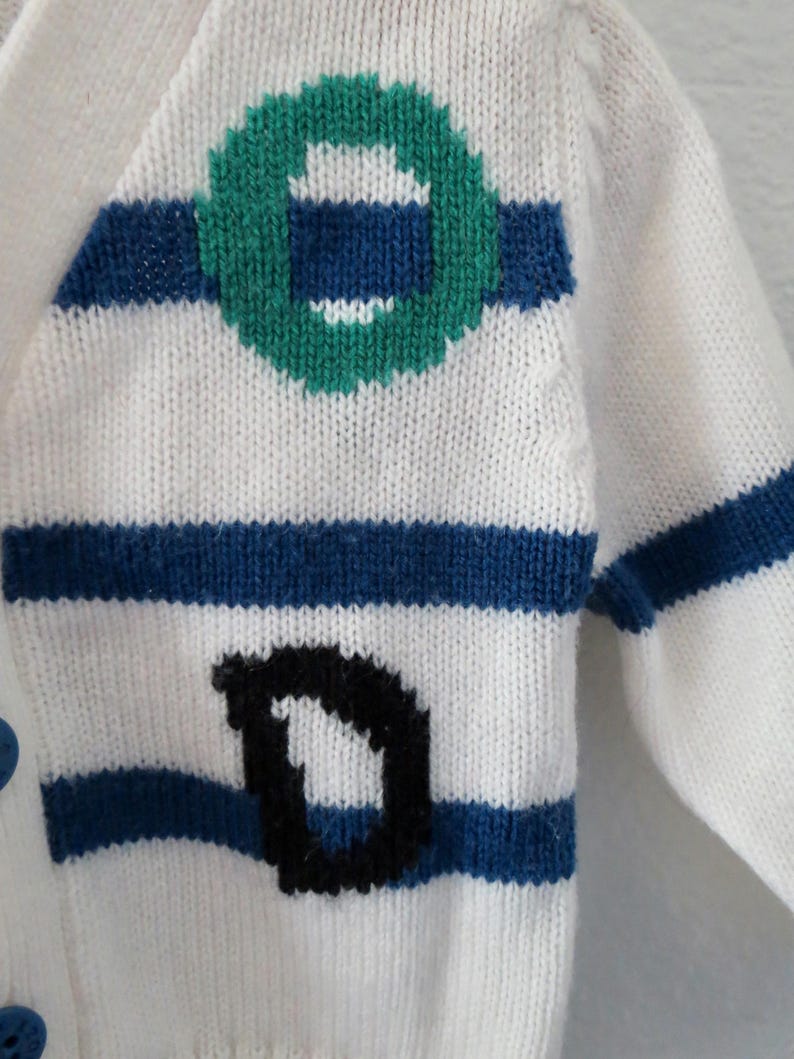 May include: A white knit cardigan featuring horizontal blue stripes, a green circular graphic, and a black letter D. The garment includes blue buttons along the front edge, showcasing a textured knit pattern throughout the fabric construction.