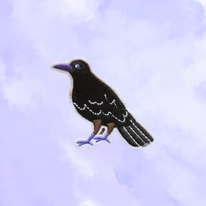 May include: Embroidered patch of a black crow with a purple beak and feet. The crow has white stitching around the edges and on the wings. The background is a light purple with a cloudy design.