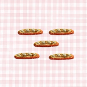 May include: Five embroidered patches shaped like baguettes. Each patch features a golden-brown crust with a textured, off-white top. The patches are arranged on a pink and white checkered background.