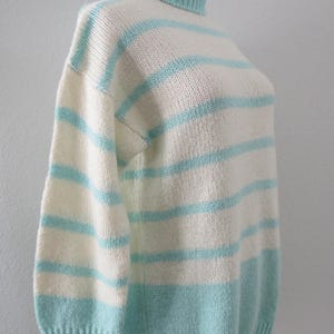 May include: A knitted sweater featuring a mock neck collar and horizontal stripes in alternating cream and light turquoise blue. The garment has long sleeves with ribbed cuffs and a ribbed hem, showcasing a consistent knit texture throughout.