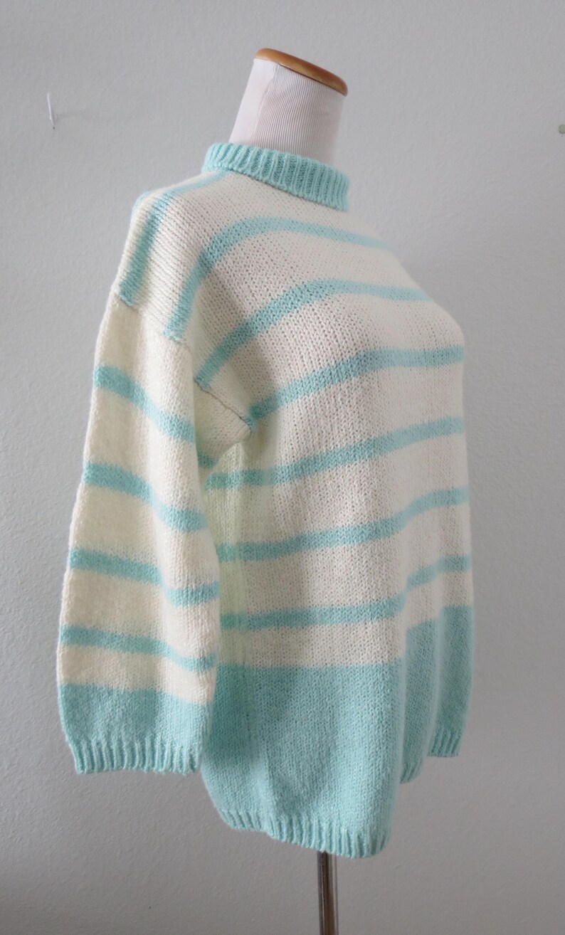 May include: A knitted sweater featuring a mock neck collar and horizontal stripes in alternating cream and light turquoise blue. The garment has long sleeves with ribbed cuffs and a ribbed hem, showcasing a consistent knit texture throughout.