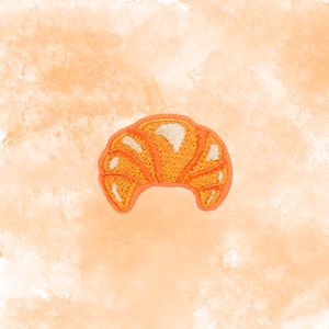 May include: Embroidered patch in the shape of a croissant. The patch features a bright orange color with white accents, and a detailed stitched design. The background is a watercolor wash of orange and peach tones.