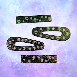 May include: Set of four black hair clips with glitter and alien and star designs. Two clips are oval shaped and two are rectangular.