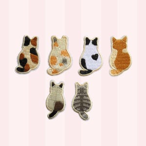 May include: Six embroidered cat patches in various coat patterns including calico, tortoiseshell, black and white with a heart shape, orange tabby, Siamese, and grey tabby. Each patch depicts a cat viewed from behind with a stitched border.