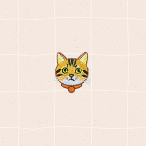 May include: Embroidered patch featuring a cat's face with yellow and black stripes, yellow eyes, and an orange collar. The patch is on a light pink background with a grid pattern. The cat's expression appears slightly sad.