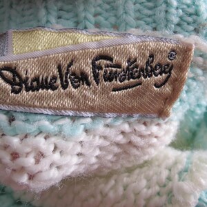 May include: A light mint green and white knit garment featuring a rectangular fabric label sewn onto the interior. The label is beige with a yellow accent and displays the black embroidered text Diane Von Furstenberg in a cursive script.