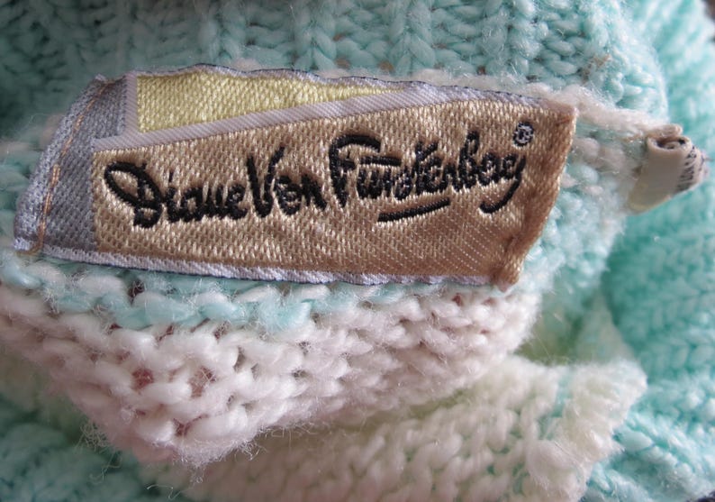 May include: A light mint green and white knit garment featuring a rectangular fabric label sewn onto the interior. The label is beige with a yellow accent and displays the black embroidered text Diane Von Furstenberg in a cursive script.
