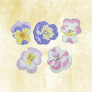 May include: Five embroidered pansy flower patches in various colors. The patches feature yellow, purple, pink, and white petals with detailed stitching. The patches are on a light yellow background.