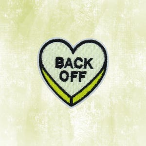 May include: A heart-shaped patch with the words "BACK OFF" in black block letters. The patch has a light green center, a black border, and a yellow accent. The background is a light green.