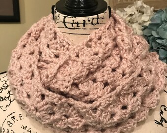May include: A blush pink crocheted infinity scarf with a delicate open stitch pattern. The scarf is displayed on a mannequin with a black stand.