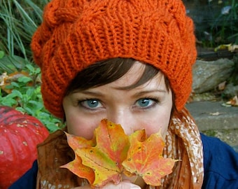May include: A woman wearing a bright orange knitted hat with a cable knit pattern, holding two autumn leaves in front of her face. She is wearing a scarf with a floral pattern and a blue shirt.