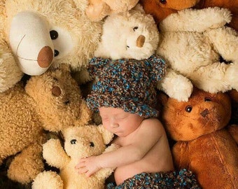 May include: A baby wearing a brown and blue crocheted hat and matching shorts is surrounded by a variety of plush teddy bears in various shades of brown and cream.