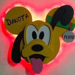 May include: A yellow and black Pluto character with a red light behind it. The character has a green collar and a tag that says "Pluto". The character's ear has the name "Dakota" written on it.