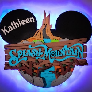 May include: A brown wooden sign with the words "Splash Mountain" in blue cursive lettering. The sign is shaped like Mickey Mouse ears with the left ear featuring the name "Kathleen" in white lettering.