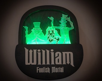 May include: A black and green light-up sign with the name "William" and the text "Foolish Mortal" underneath. The sign features three ghostly figures from the Haunted Mansion attraction.