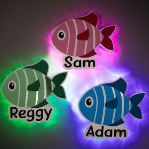 May include: Three light-up fish with names written below them. The fish are pink, green, and blue with white stripes. The names are Sam, Reggie, and Adam.