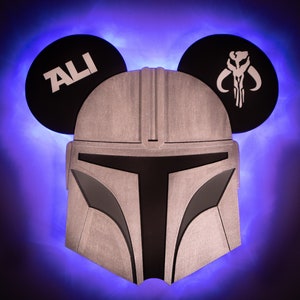 May include: A silver Mandalorian helmet with a black visor, decorated with two black Mickey Mouse ears. One ear has the word "ALI" written on it, and the other ear has a stylized Mandalorian symbol.