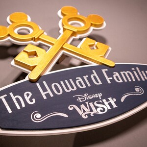 May include: A wooden sign featuring the text "The Howard Family" and the Disney Wish logo. The sign is navy blue with white trim and silver lettering. Two gold-colored keys with Mickey Mouse silhouettes are crossed above the sign.