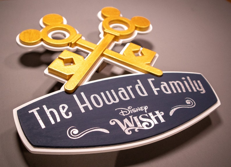 May include: A wooden sign featuring the text "The Howard Family" and the Disney Wish logo. The sign is navy blue with white trim and silver lettering. Two gold-colored keys with Mickey Mouse silhouettes are crossed above the sign.