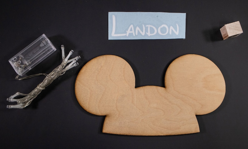 May include: A wooden Mickey Mouse-shaped sign with a light string and a battery pack. The sign is blank and ready to be personalized. The name Landon is written on a blue piece of paper.