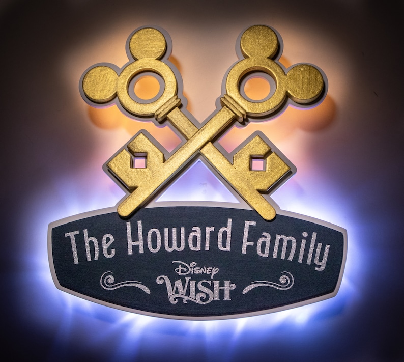 May include: A custom family sign featuring crossed gold keys with Mickey Mouse ear designs. The sign includes the text "The Howard Family" and "Disney Wish" in silver lettering on a dark blue background, with a glowing blue border.
