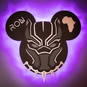 May include: Black Panther mask with a silver collar, in the shape of Mickey Mouse ears, with the word "RON" on the left ear and a silhouette of Africa on the right ear. The background is a purple glow.
