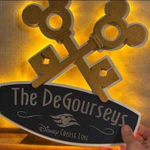 May include: A decorative sign featuring two crossed gold keys with Mickey Mouse ear designs. The sign has a black oval base with the text "The DeGourseys" and "Disney Cruise Line" in silver glitter.