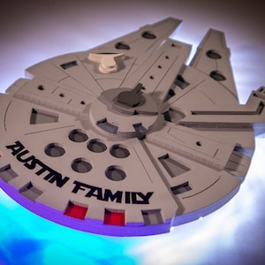 Könnte beinhalten: Ein weißes, 3D-gedrucktes Modell des Raumschiffs Millennium Falcon aus Star Wars. Das Modell ist mit den Worten "Austin Family" in schwarzer Schrift verziert.
