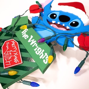 May include: A blue cartoon character wearing a Santa hat and red gloves, with a green Christmas tree and string lights. The tree has a red tag that says "Merry Xmas From Stitch". The green sign says "the Wrights".