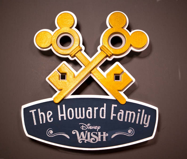 May include: A wooden sign featuring two crossed gold keys with Mickey Mouse-shaped heads. The sign includes the text "The Howard Family" above "Disney Wish" in a decorative font, set against a navy blue background with a white border.