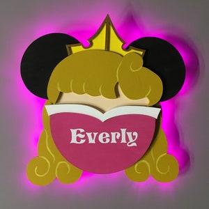 May include: A pink and yellow princess-themed nightlight with the name "Everly" written on it. The nightlight has a crown, mouse ears, and a pink heart-shaped face.