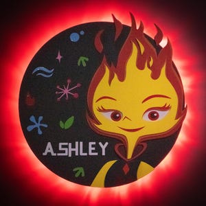 May include: A circular design featuring a cartoon character with a flame-like head, yellow face, and the name "ASHLEY" in white. The design includes water, air, and earth symbols, set against a dark background with a red glowing border.