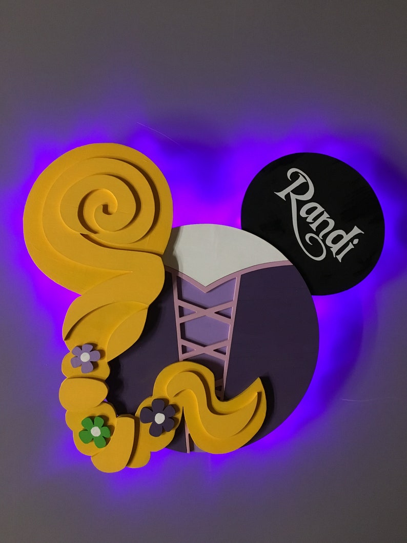 May include: Purple and yellow Rapunzel-themed Mickey Mouse ears with the name "Randi" written in white on the black ear.