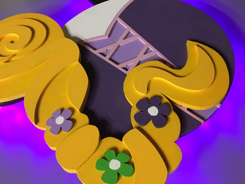 May include: A purple and yellow wooden wall decor featuring a stylized image of a woman's hair with flowers. The hair is yellow and the flowers are purple, green, and white.